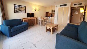 A compact living space with two sofas, a small dining table, a TV, and a kitchenette.
