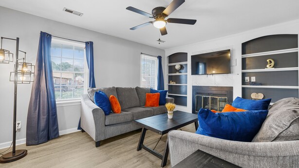 A living room with a gray sofa, blue and orange cushions, a coffee table, a fireplace, and a ceiling fan. - The Full House (Chesapeake)
