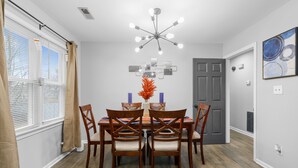 A dining area with a wooden table and chairs, a modern chandelier, and a vase with orange flowers. - The Full House (Chesapeake)