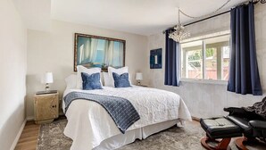 Ein Schlafzimmer mit einem Bett, einem Sessel, einem Nachttisch, einem Gemälde und einem Fenster mit Vorhängen.