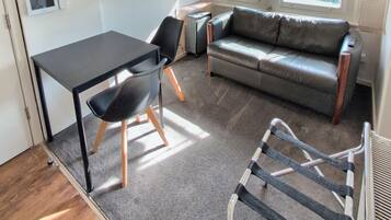 A compact living space with a sofa, a dining table, a chair, a radiator, and a window with blinds.