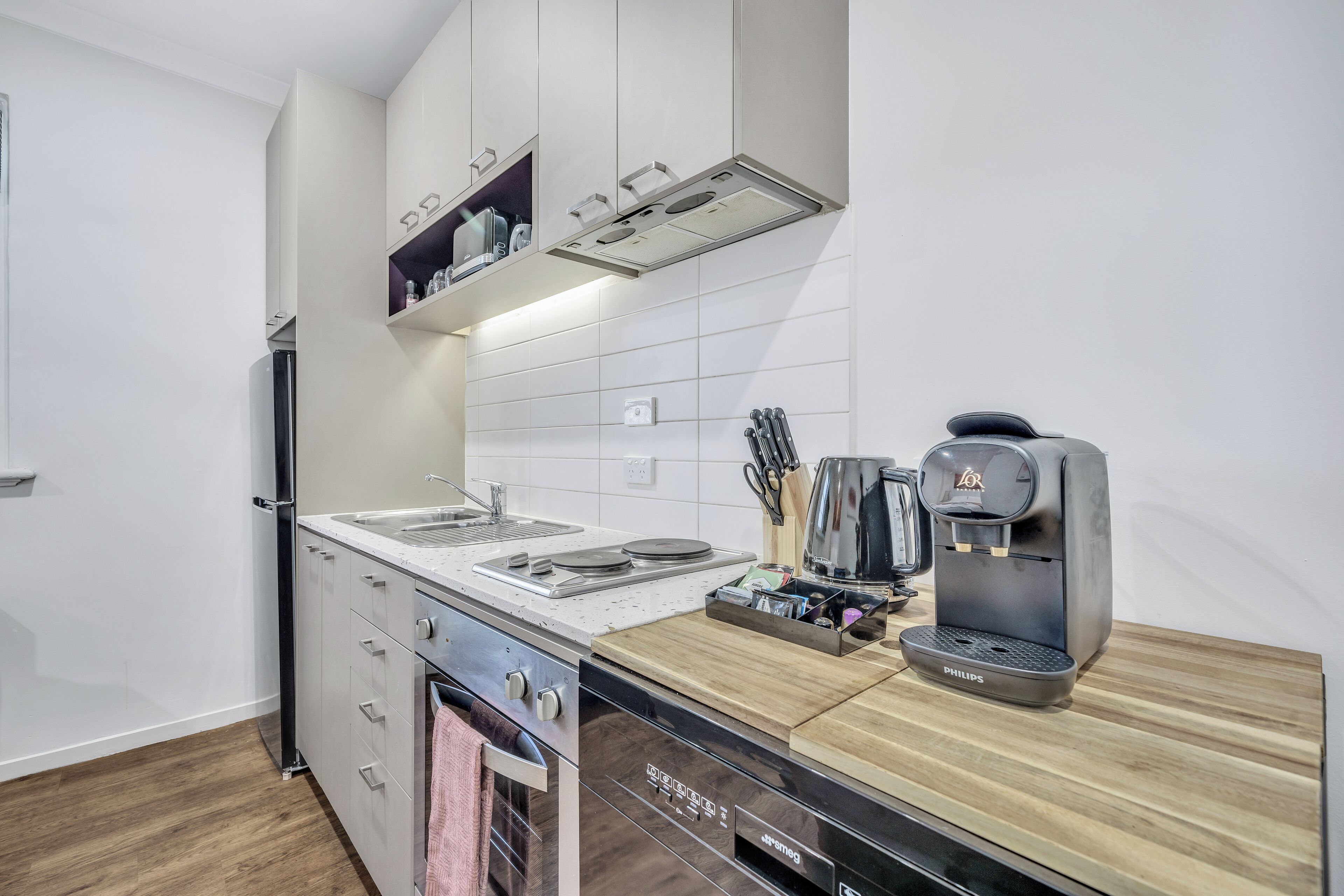 A modern kitchen with white cabinets, a built-in oven, a dishwasher, a coffee maker, and a wooden countertop.