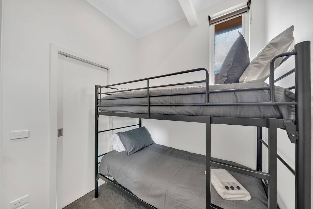 A bunk bed with grey bedding and pillows, a white door, and a window with blinds.