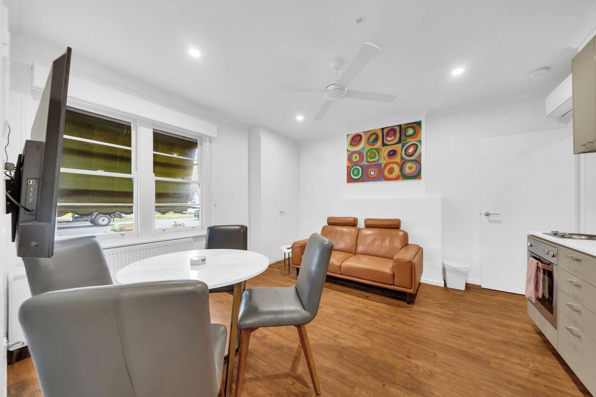 A modern living room with a brown leather sofa, a round dining table with chairs, a ceiling fan, and a window with blinds.