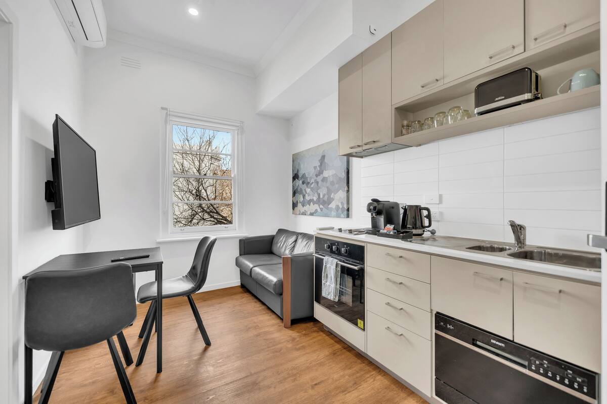 A modern kitchen with a built-in oven, a coffee maker, and a dishwasher. There is a dining area with a table and chairs, and a gray sofa.