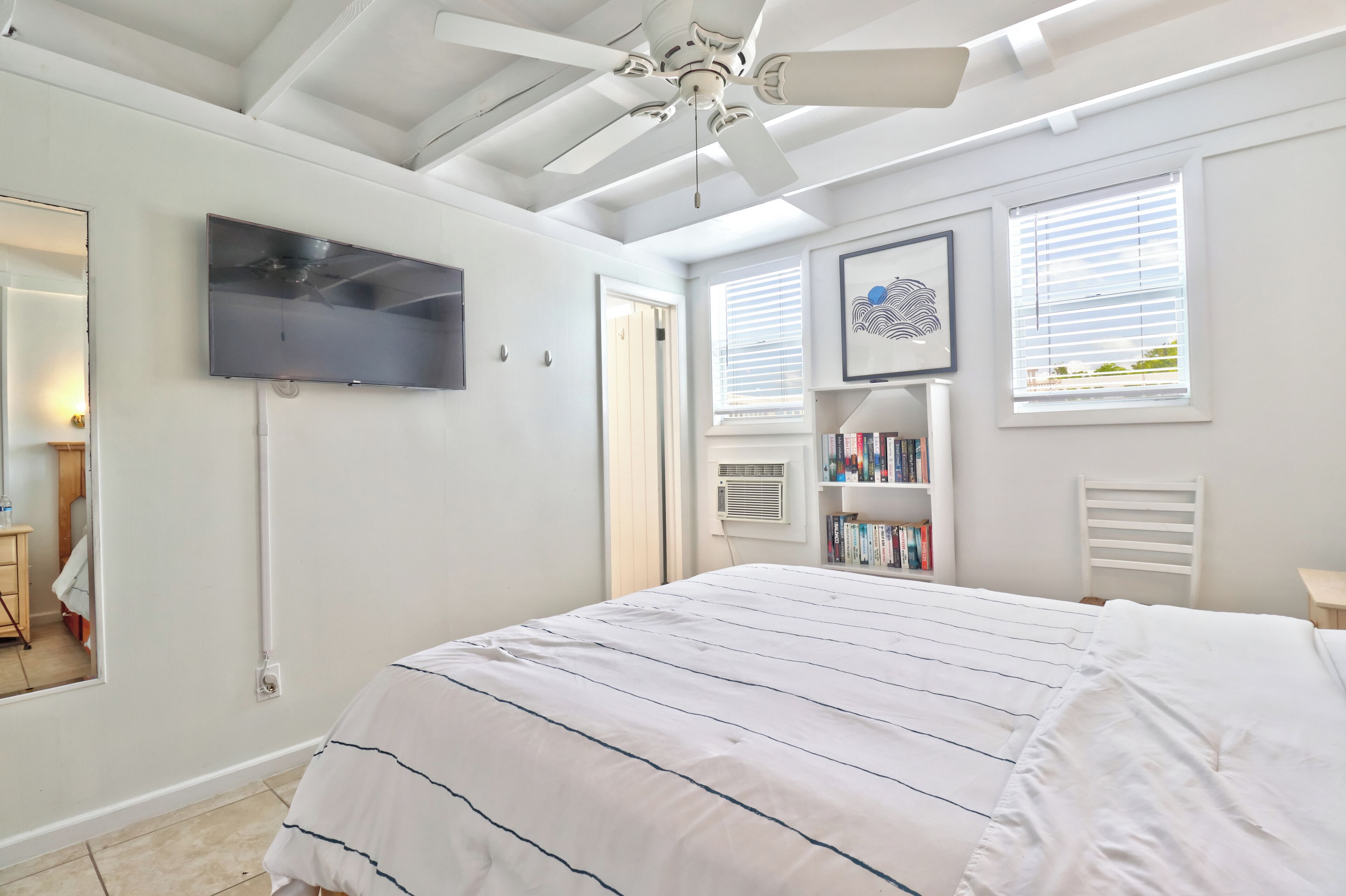 Une chambre à coucher comprenant un lit, un ventilateur de plafond, un téléviseur fixé au mur, une bibliothèque et une fenêtre avec des stores.
