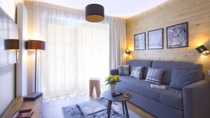 Una sala de estar moderna con un sofá gris, una mesita redonda con un florero, y un ventanal con cortinas traslúcidas.