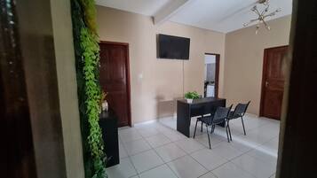 Uma sala de jantar com mesa e cadeiras, uma televisão fixada na parede e um ventilador de teto.