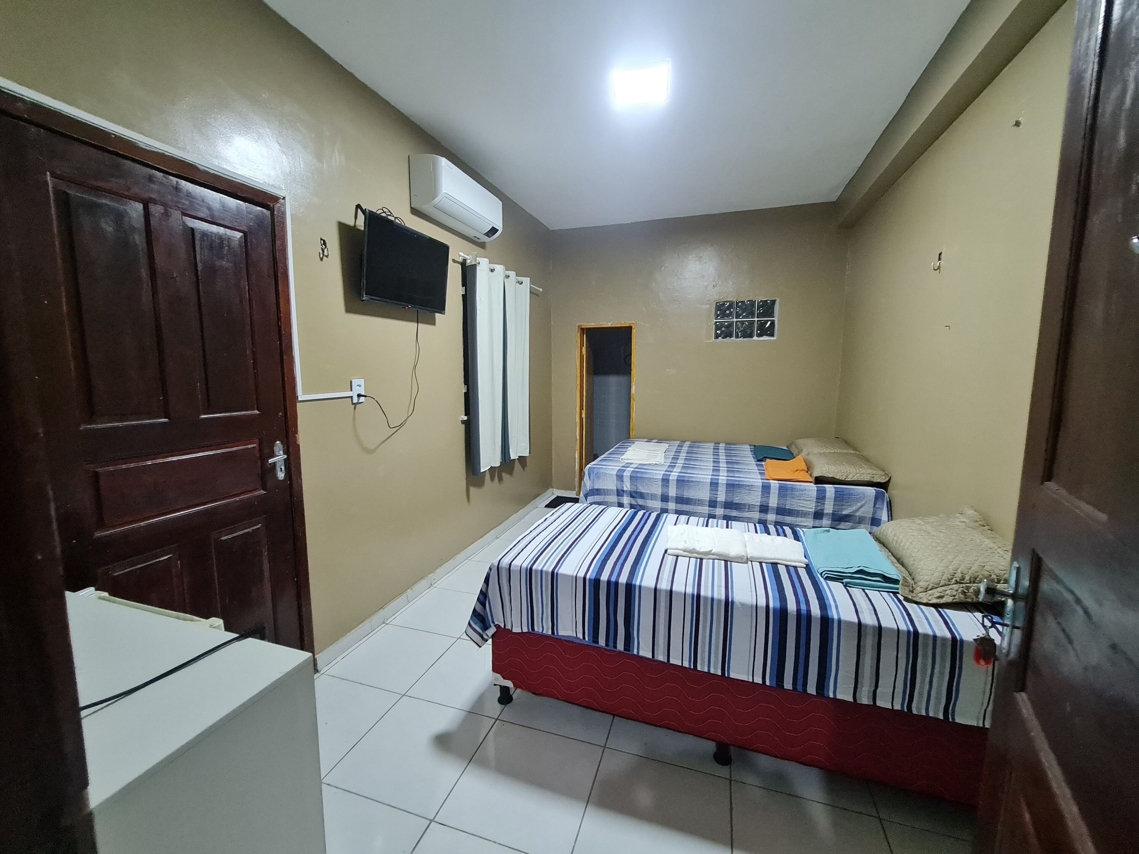 Quarto pequeno e bem iluminado com duas camas, televisão e frigorífico.