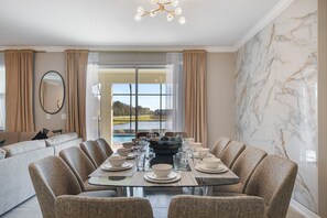 A dining area with a glass table, beige upholstered chairs, and a view of a lake through large windows.