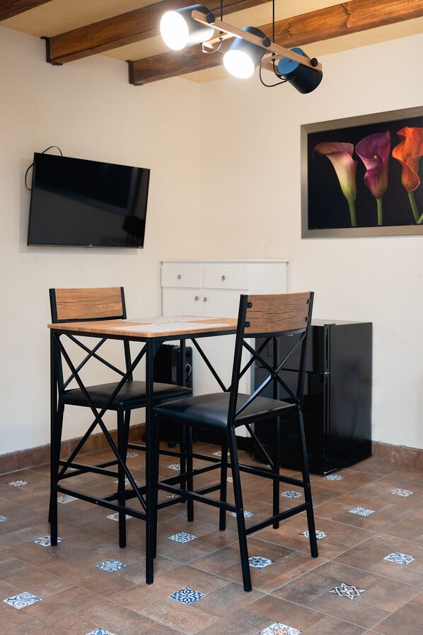 A dining area with a table and chairs, a television mounted on the wall, and a refrigerator.