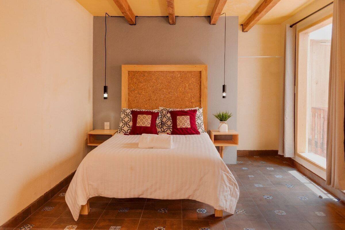 Una cama bien hecha con almohadillas estampadas, un cabecero de madera y una pequeña planta en una mesita de noche.