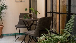 Un pequeño patio con mesa y dos sillas, rodeado de plantas en macetas y un ventanal con vistas.
