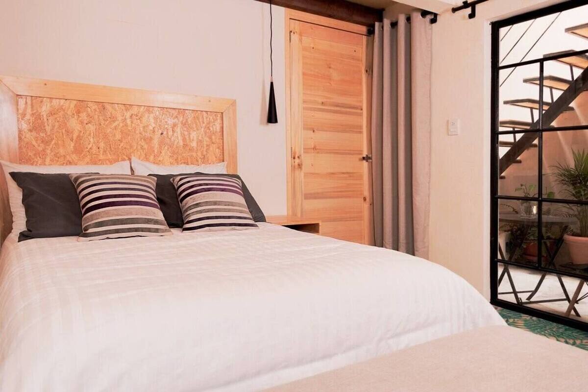Un dormitorio con cabecera de madera, cama con ropa de cama blanca, almohadas a rayas, y vistas de una escalera y plantas a través de una puerta de cristal.
