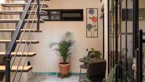 Una escalera con una planta en maceta a su lado, una pequeña mesa con una silla, y una ventana con un cuadro de flores.