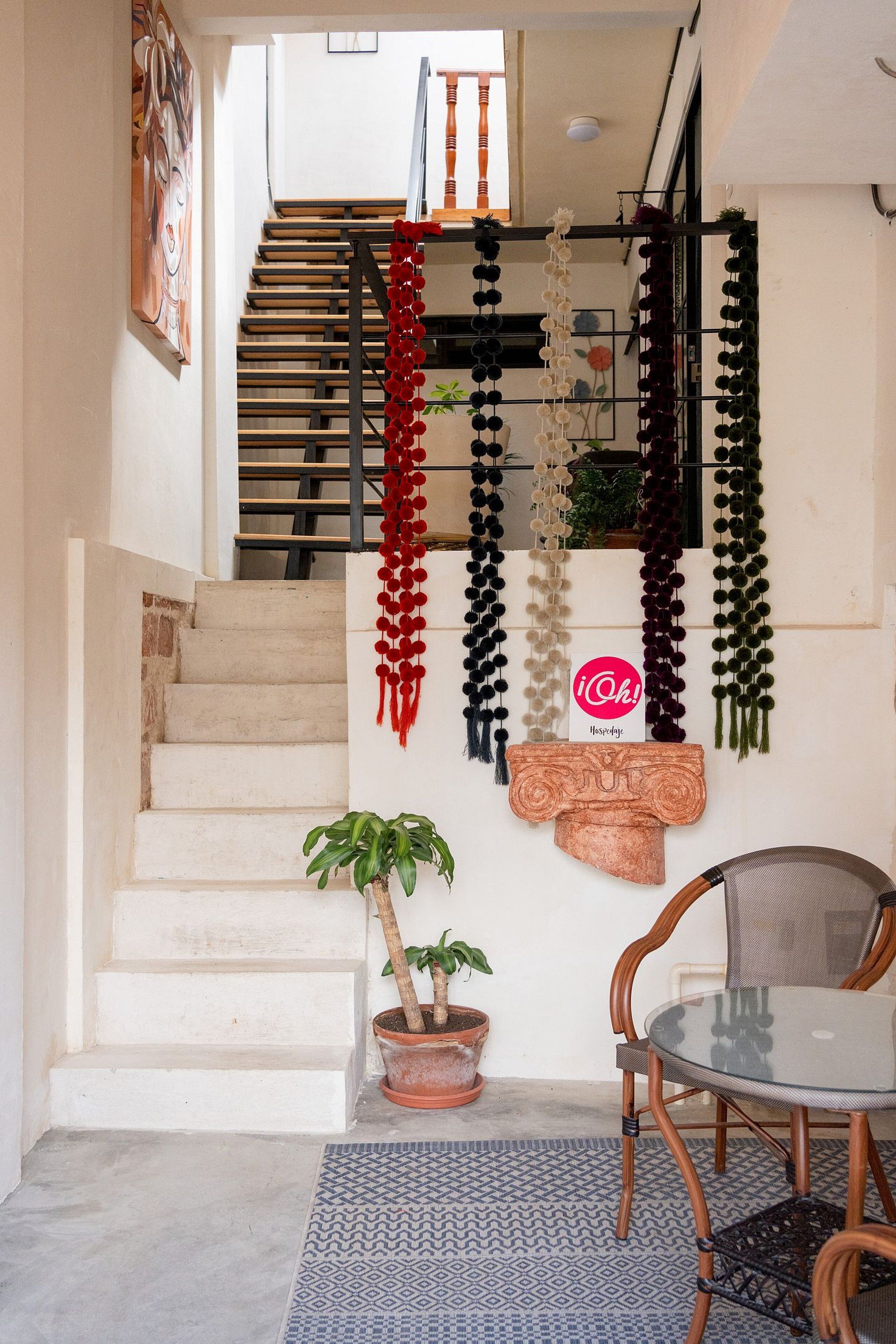 A staircase with a hanging display of various items, a potted plant, a round glass table with chairs, and a patterned rug.
