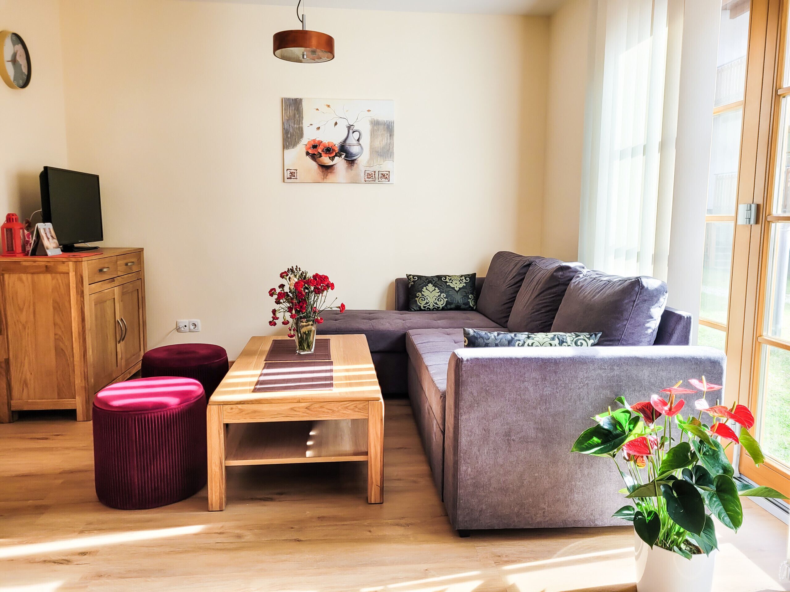 A living room with a wooden table, a grey sofa, a red ottoman, and a potted plant.