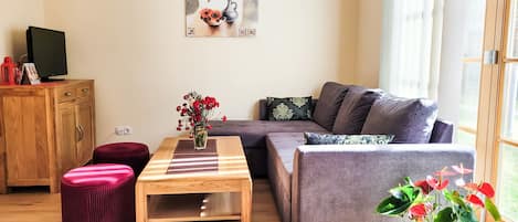 A living room with a wooden table, a grey sofa, a red ottoman, and a potted plant.