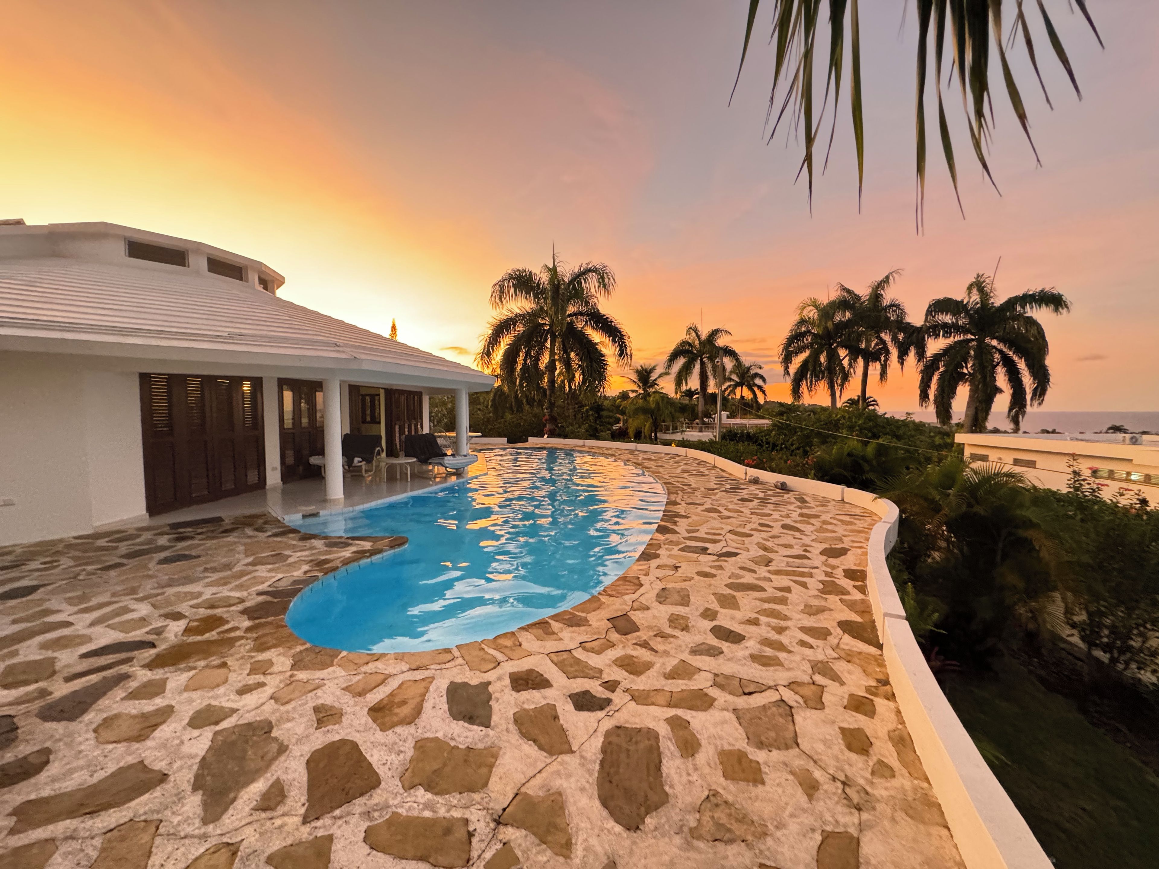 Ein Pool mit einer Steindeckung, einer überdachten Sitzgelegenheit und Palmen im Hintergrund.