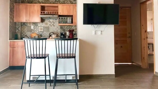 A modern kitchen with wooden cabinets, a tiled backsplash, and a flat-screen TV mounted on the wall. There are two black bar stools at a small island. - Oh Baja California (San Cristóbal de las Casas)