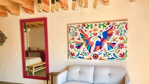 A living room with a light grey sofa, a colorful floral painting, a mirror with a red frame, and wooden ceiling beams. - Oh Baja California (San Cristóbal de las Casas)