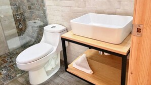 A bathroom with a toilet, a white sink on a wooden shelf, and a shower area with patterned tiles. - Oh Baja California (San Cristóbal de las Casas)