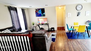 Una sala de estar con un sofá, un televisor sobre un mueble, una mesa de comedor con sillas y un reloj en la pared.