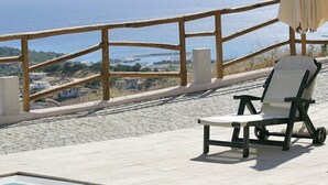 A poolside area with a wooden railing, a white lounge chair, and a view of the sea and buildings in the background. - Hotel Castello Budoni (Budoni)