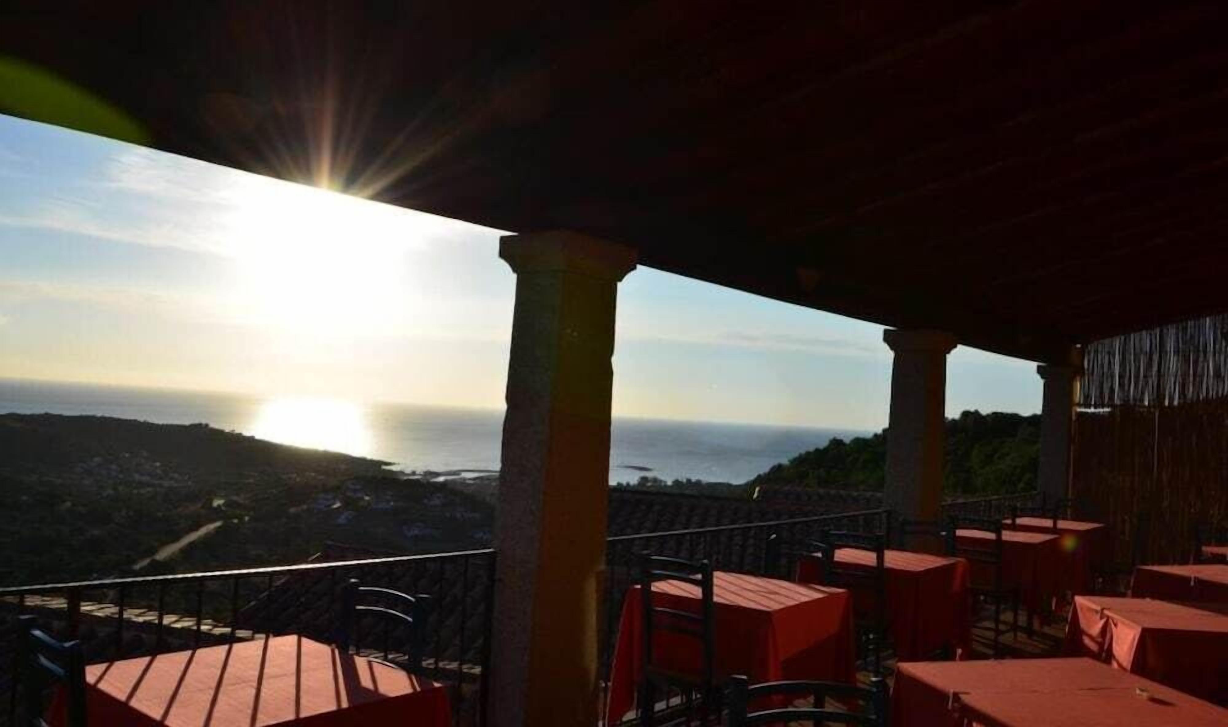 A terrace with tables and chairs, overlooking a coastal landscape with a sunset.