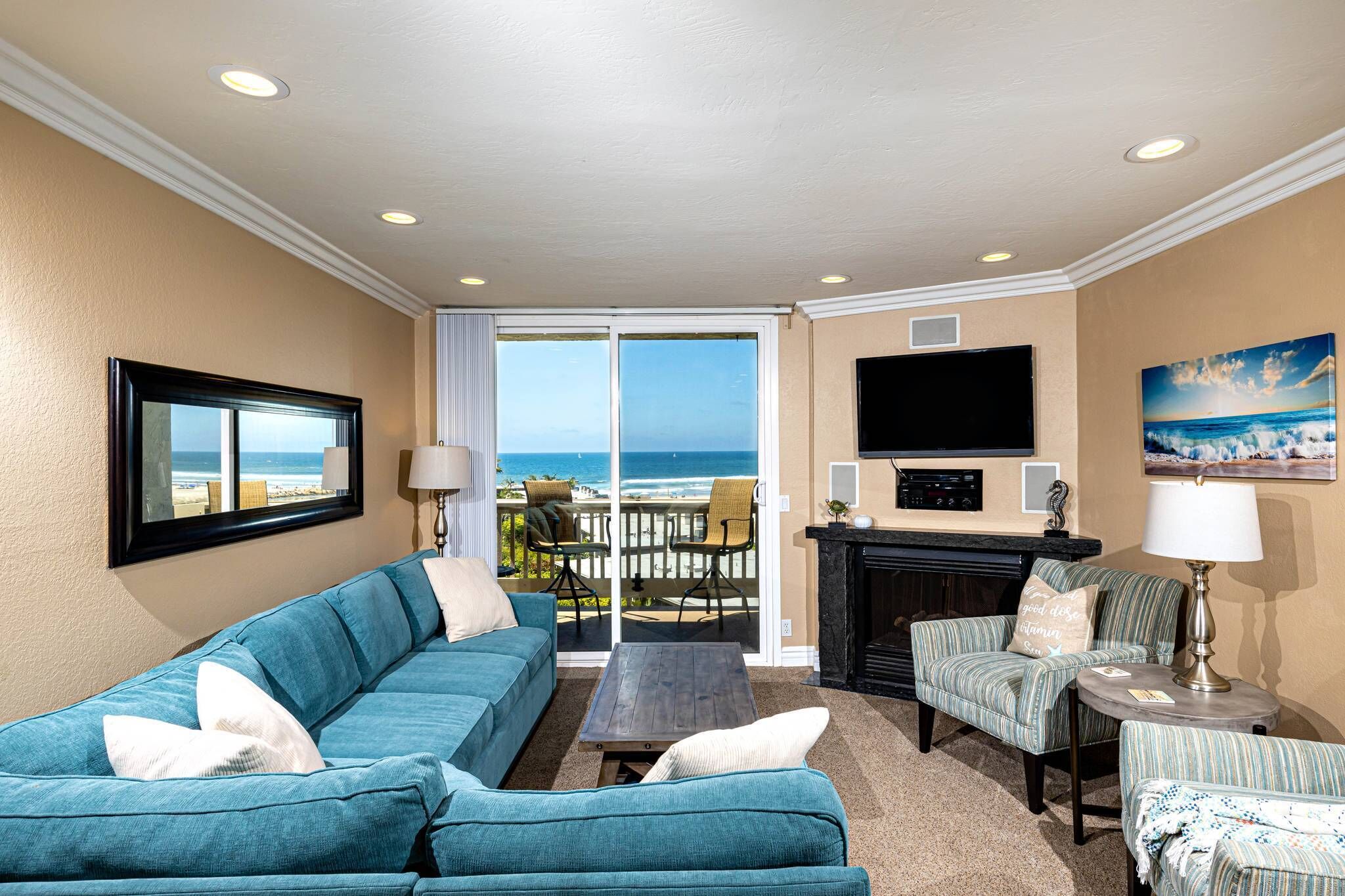 A living room with a blue sofa, a coffee table, a fireplace, and a TV. There is a balcony with a view of the ocean.