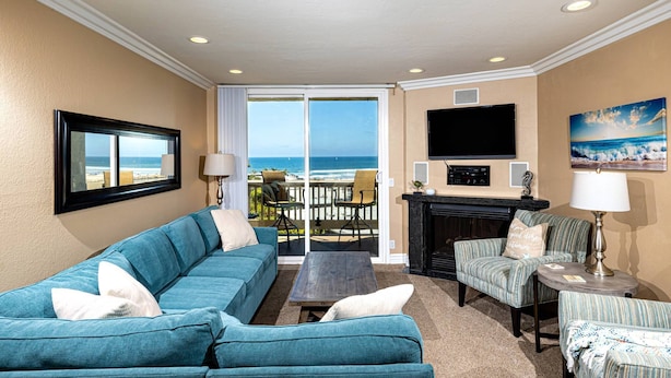 A living room with a blue sofa, a coffee table, a fireplace, and a TV. There is a balcony with a view of the ocean. - North Coast Village 3brm Penthouse - Amazing Oceanfront Panoramic Views -  G315 (Oceanside)