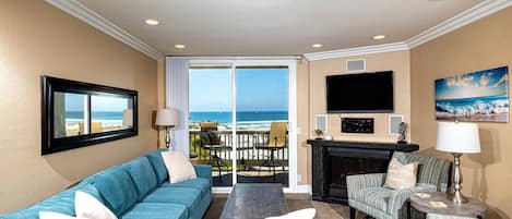A living room with a blue sofa, a coffee table, a fireplace, and a TV. There is a balcony with a view of the ocean.