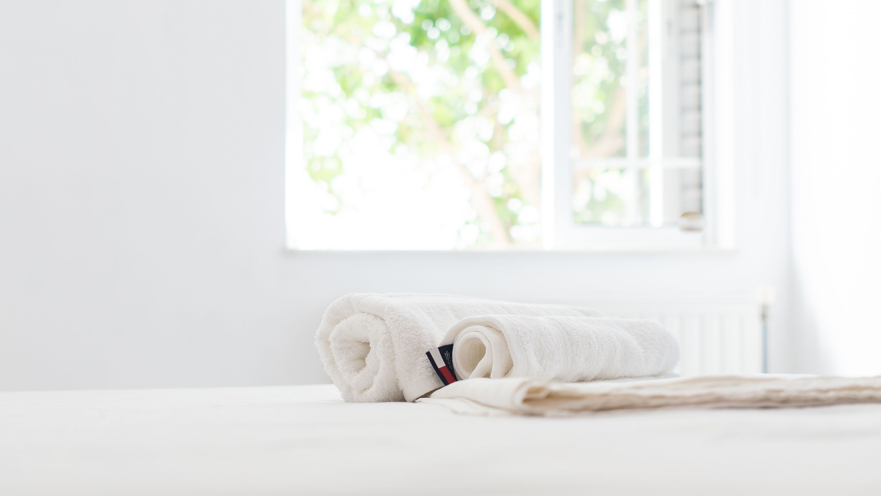 Une serviette blanche, soigneusement pliée, est posée sur un lit, avec une fenêtre en arrière-plan.