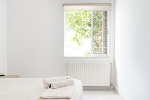 Une chambre à coucher au style minimaliste, avec une grande fenêtre, un radiateur et un lit soigneusement fait, sur lequel est posée une serviette pliée.