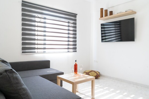 Un salon moderne avec un canapé gris, une petite table en bois sur laquelle se trouvent une bouteille et des verres, une grande fenêtre avec des stores, un téléviseur fixé au mur et une étagère ornée d’objets décoratifs.