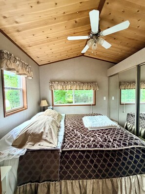 A bedroom with a wooden ceiling, a bed with patterned bedding, a ceiling fan, and windows with curtains.