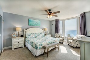 Ein Schlafzimmer mit einem Bett, zwei Nachttischen, einer Bank, einem Sessel, einem Schreibtisch, einem Deckenventilator und einem großen Fenster mit Vorhängen.