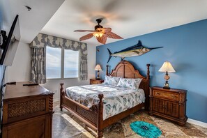 Une chambre avec un lit en bois, deux tables de chevet, un ventilateur de plafond, une grande fresque représentant des poissons et une vue sur la mer par la fenêtre.