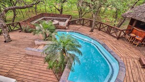 A wooden deck with a swimming pool, surrounded by trees and a thatched-roof structure.