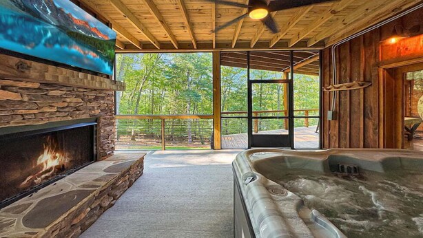 Ein gemütliches Blockhaus-Interieur mit Kamin aus Stein, einem Whirlpool und großen Fenstern mit Blick auf den Wald.