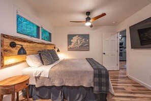 Ein Schlafzimmer mit einem hölzernen Kopfende, einem Bett mit grau-weißer Bettdecke, einem Nachttisch und einem Deckenventilator.