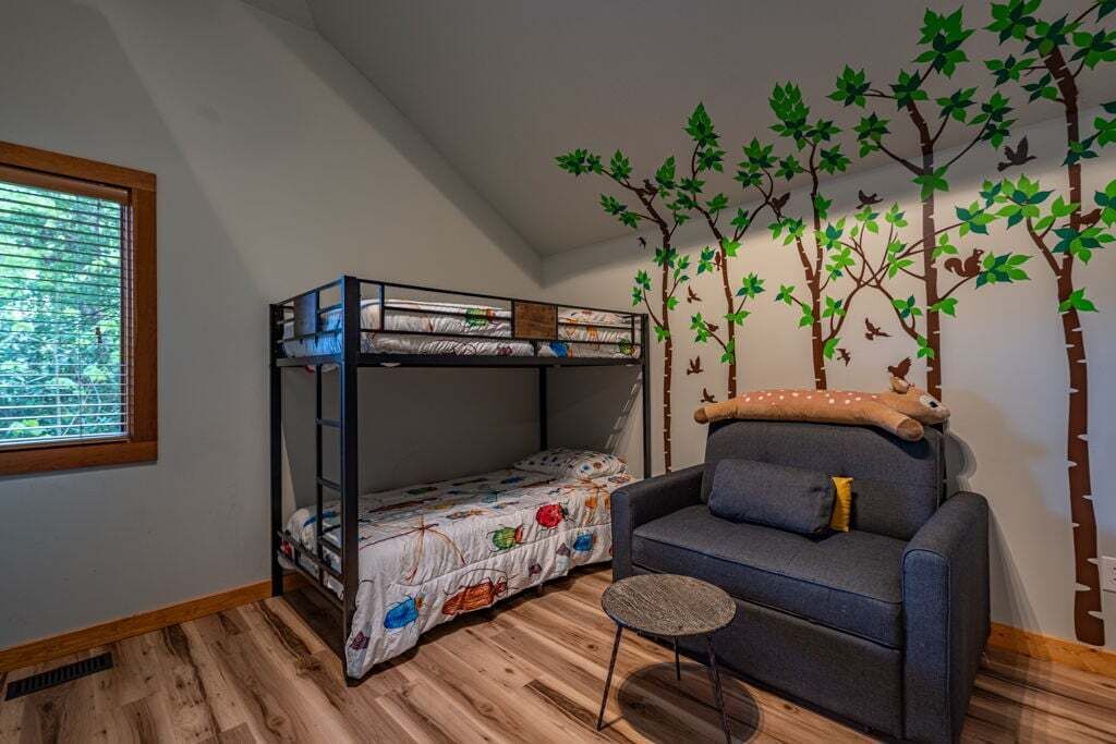 Quarto de criança com beliche, sofá, mesa redonda pequena e um mural de árvore na parede.