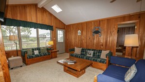 A cozy cabin interior with wooden walls, a blue sofa, a green plaid sofa, a wooden coffee table, and a small table with a lamp. - Hameljuice - Classic 3 BR Oceanside Beach Cottage - close to beach (Southern Shores)