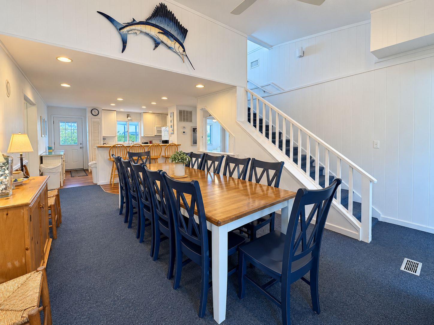 A spacious dining area with a long wooden table and blue chairs, a kitchen in the background, and a staircase leading to an upper level.