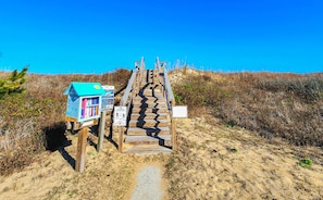 Un escalier en bois mène à une zone de plage où se trouve une petite structure bleue et des panneaux d’information.