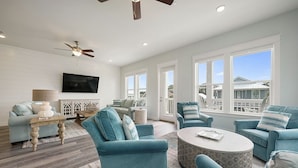 A spacious living room with teal furniture, a ceiling fan, and large windows offering a view of the neighborhood. - Dusk Til Dune - Vacation Home Near the Beach with Shared Pool (Santa Rosa Beach)