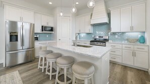 A modern kitchen with white cabinets, stainless steel appliances, a central island with stools, and pendant lighting. - Dusk Til Dune - Vacation Home Near the Beach with Shared Pool (Santa Rosa Beach)