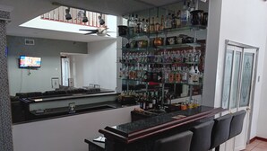 Una zona de bar con mostrador, varias estanterías llenas de botellas de licor y tres taburetes.