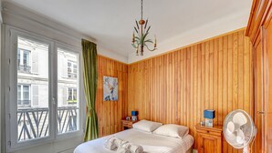 A bedroom with a large bed, wooden paneling, a chandelier, a fan, and a view of buildings outside.