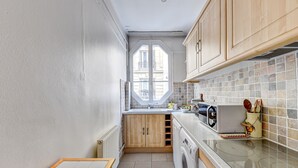 A compact kitchen with wooden cabinets, a built-in oven, a washing machine, and a window with a view of buildings.